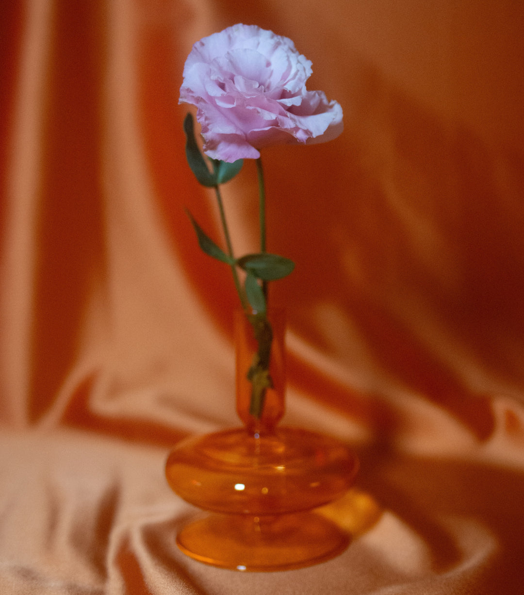 Light purple flower in an orange vase with an orange silk background.