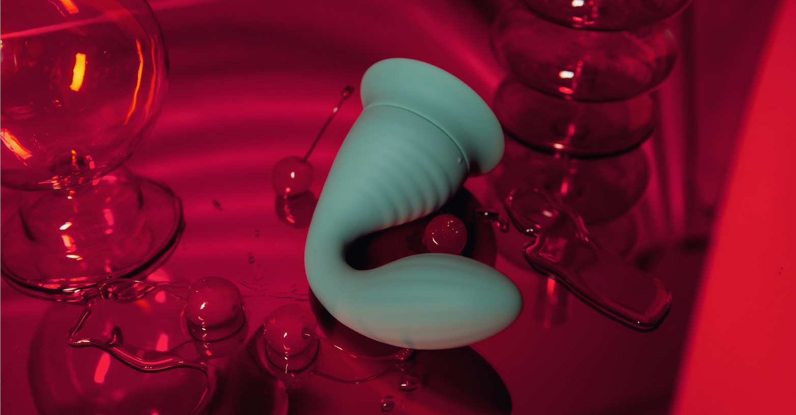 Photograph of the Dollop vibrator on a pink reflective surface surrounded by cherries.