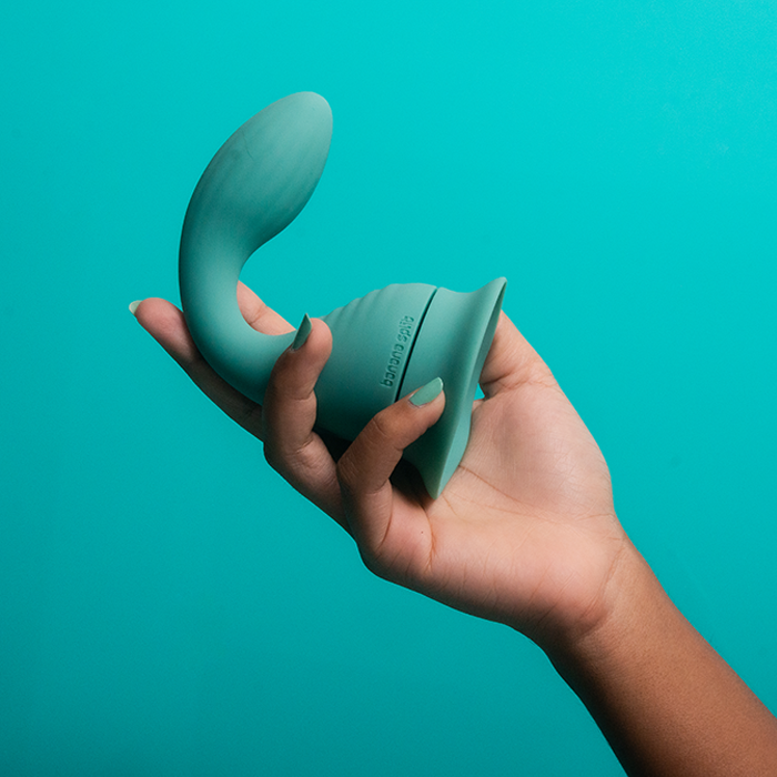 Photography of the blue Dollop vibrator connected to the suction cup held up by a woman's hand with turquoise nail polish. The Banana Split logo is debossed on the side of the vibrator. 