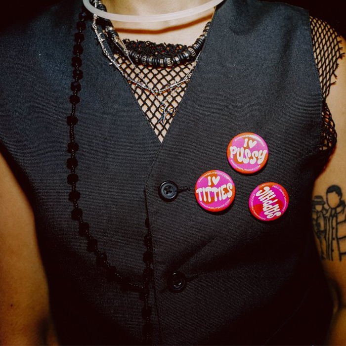 A person with a black vest wearing the set of 3 Pink and Orange Banana Split Pins that say "Sapphic," "I Heart Titties," and "I heart Pussy" as well as our website URL.